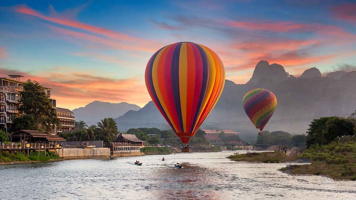 Colorful hot air balloons flying over a river at sunset in Vang Vieng, Laos, with scenic mountains in the background.