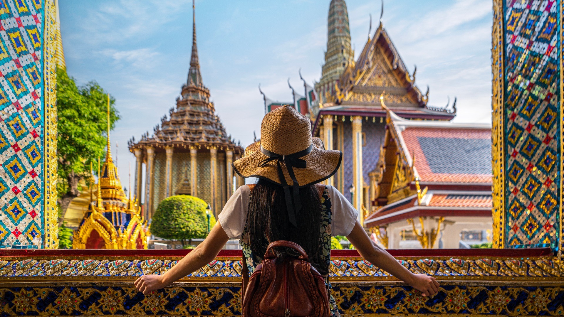A traveler wearing a straw hat and backpack admiring the intricate architecture and golden spires of a traditional temple in Thailand, showcasing the rich cultural heritage and vibrant colors typical of Thailand tours