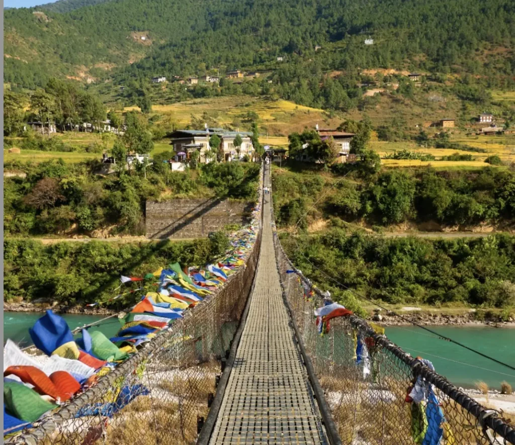 Best Things to Do in Bhutan featuring a scenic suspension bridge decorated with colorful prayer flags, stretching over a turquoise river toward a peaceful village surrounded by lush green hills.